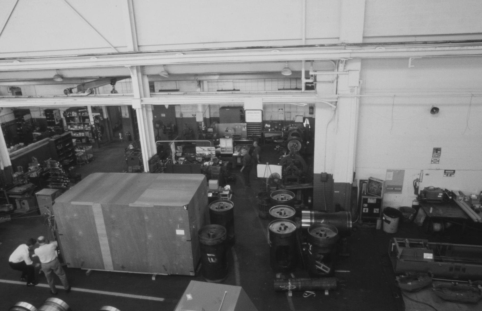 Wide elevated view looking down a machine shop aisle lined with lathes and drill presses, two men moving a wooden crate
