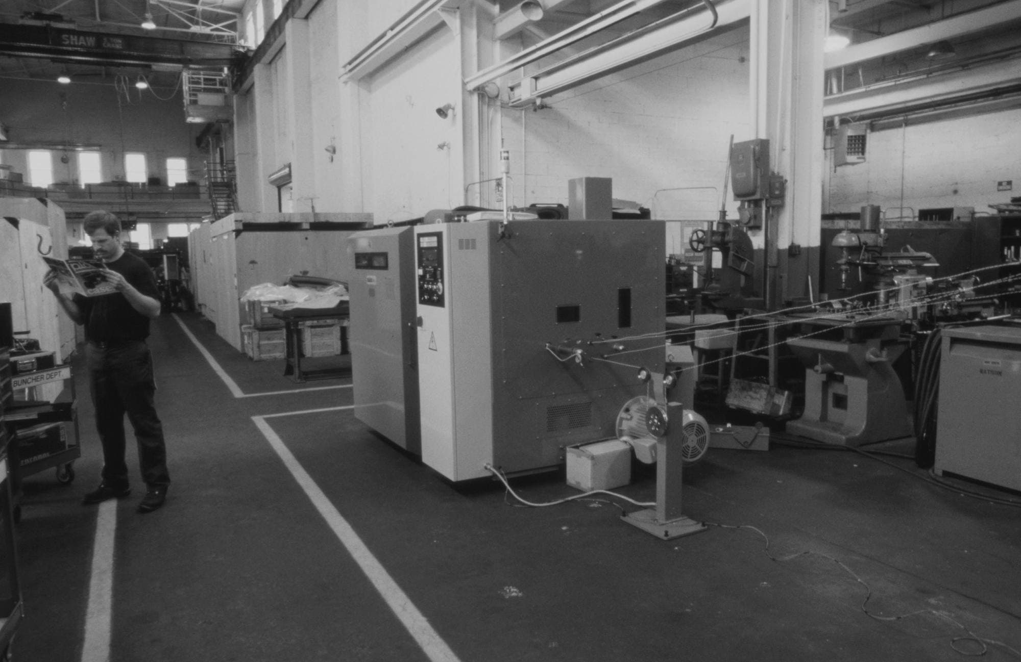 Wide view down a factory aisle with an overhead crane rail, worker reading a clipboard beside rows of machine tools