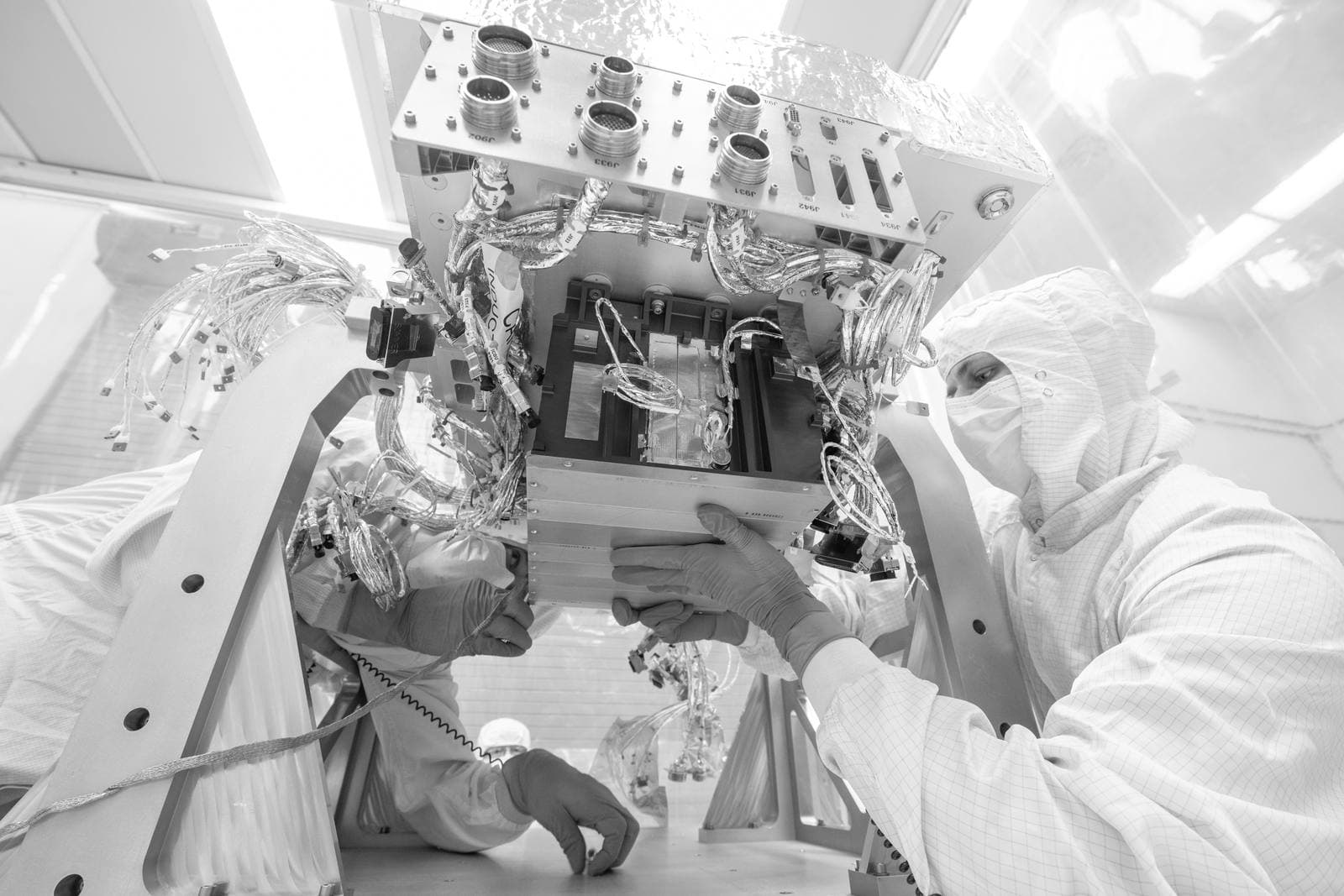 Two technicians in cleanroom suits and blue gloves lowering an electronics box, wrapped in wiring harnesses, onto a spacecraft deck