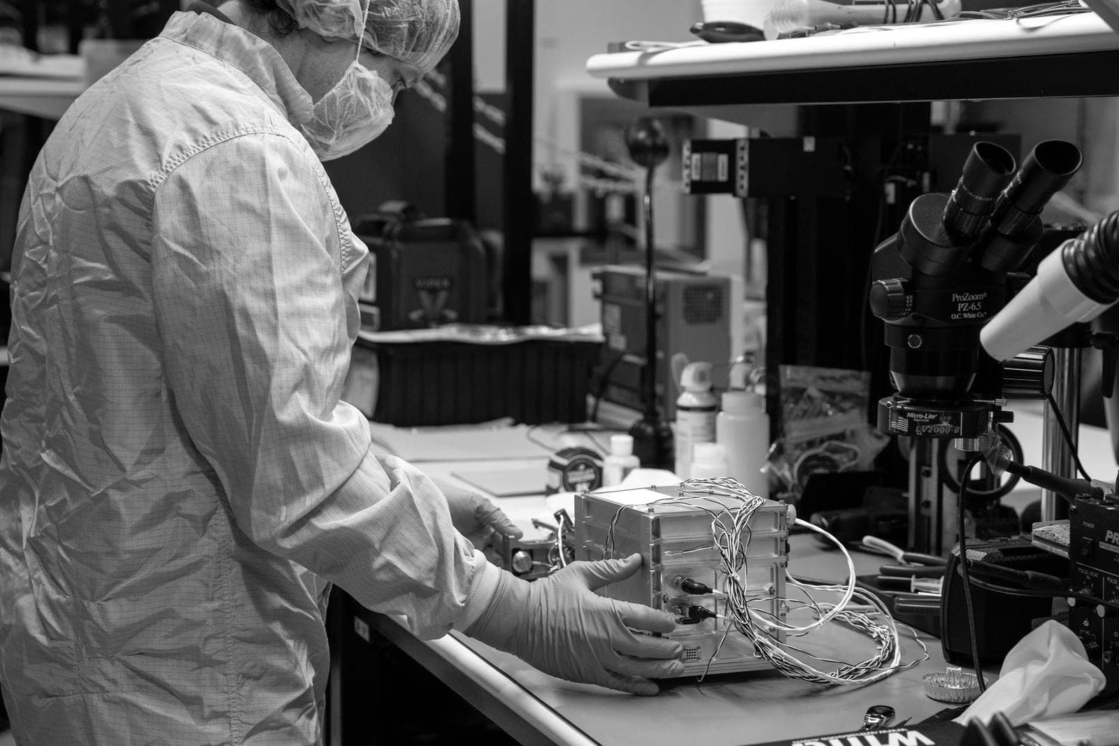 Technician in a cleanroom smock and blue gloves assembling a small instrument box at a bench fitted with a binocular microscope