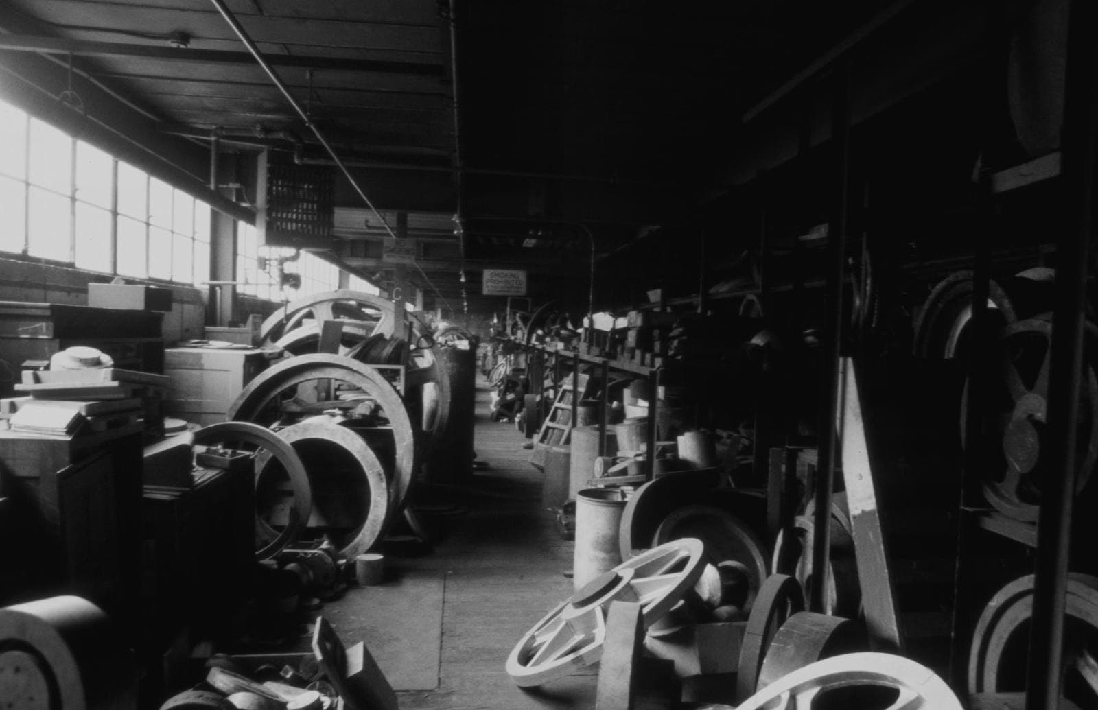 Rows of old wooden foundry patterns and cast metal wheels stored on shelving above a dim machine shop aisle