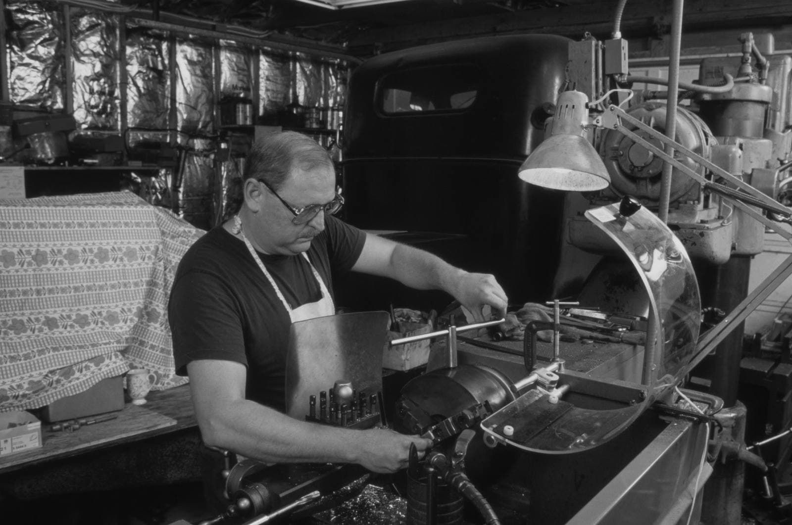 Machinist adjusting a small lathe in a home garage workshop, vintage truck cab visible behind the bench