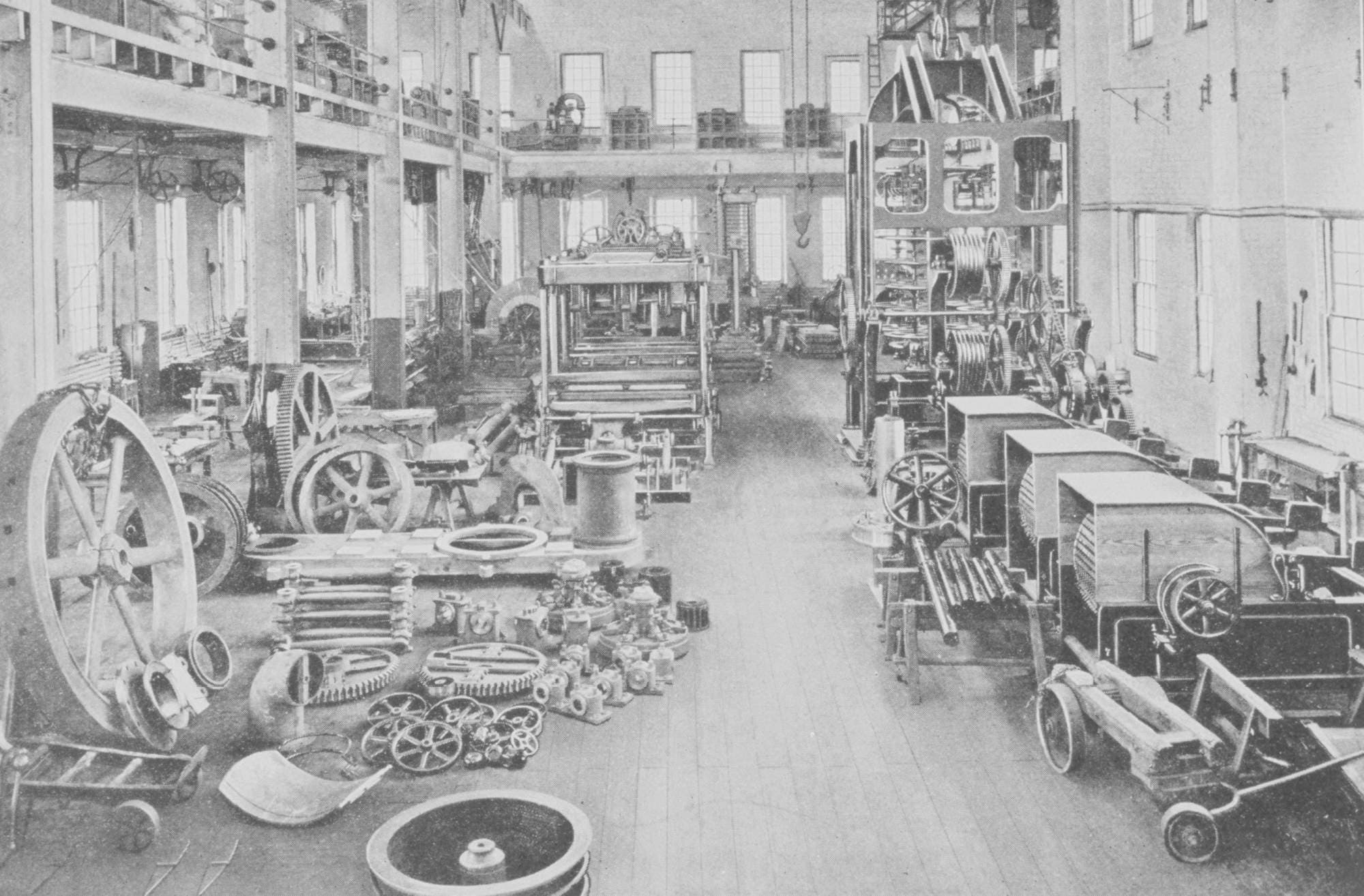 Archival photo of a two-story machine shop floor filled with belt-driven line-shaft machinery, flywheels, and gears laid out on the floor